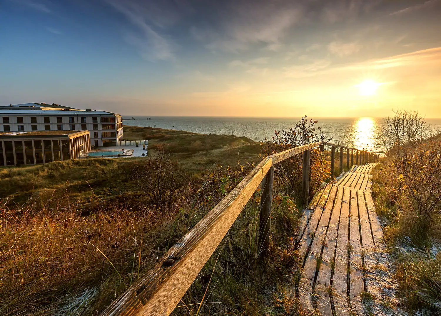 A-ROSA Sylt Una passerella di legno conduce alla spiaggia, con un edificio e lo specchio d’acqua sullo sfondo.
